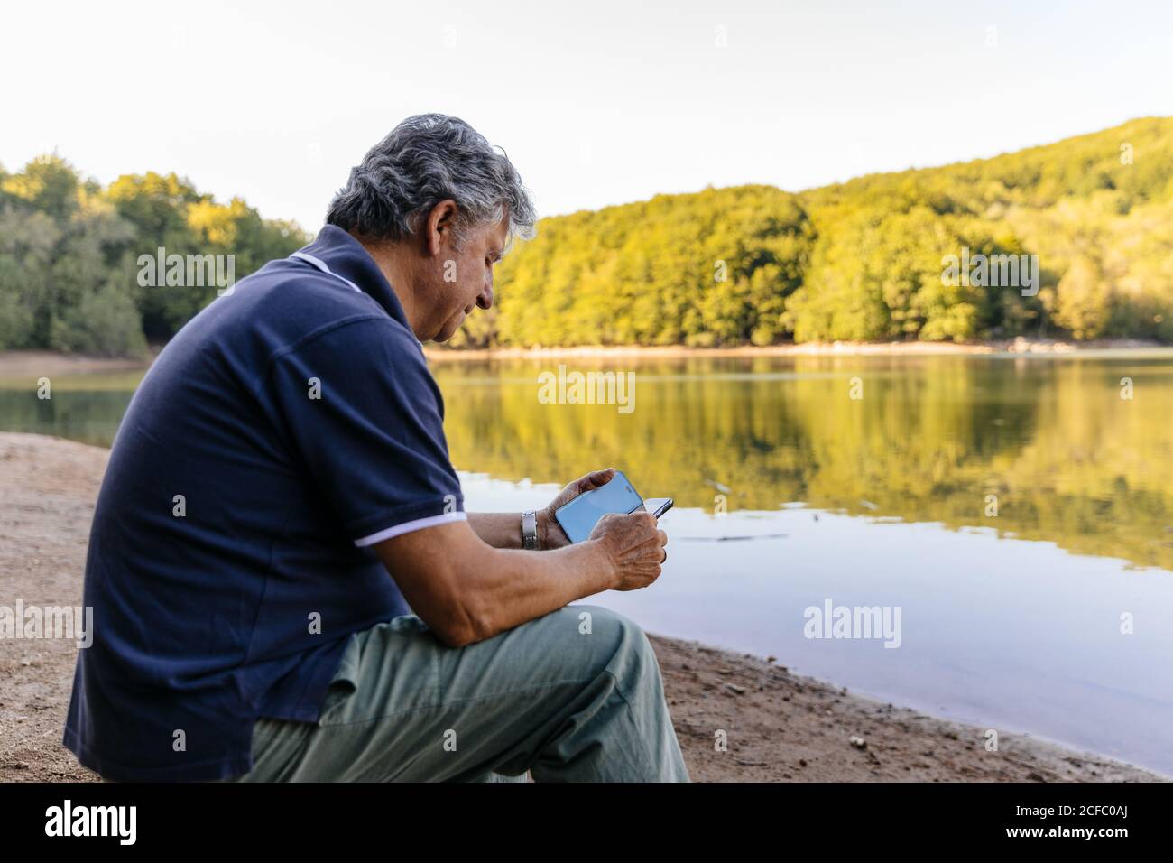 Side view of senior man using mobile phone while sitting amazing lake ...