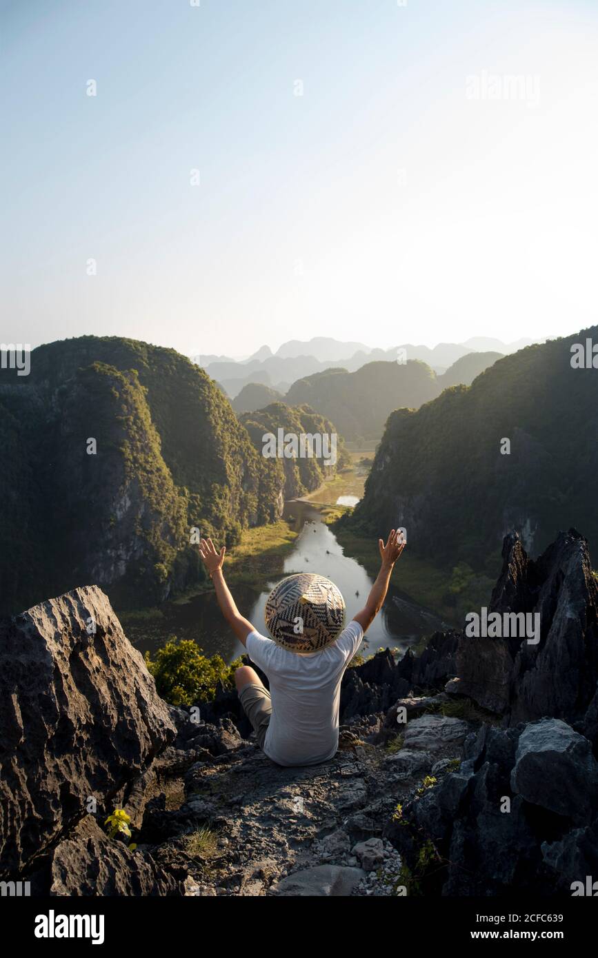 From above back view of anonymous male tourist with open arms on rocky terrain in Mua cave and observing spectacular scenery during vacation Stock Photo