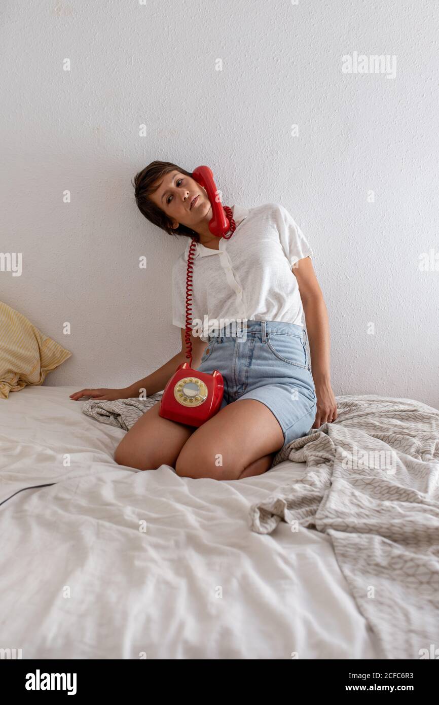 Indifferent lady with handset of red telephone near ear listening to dialogue partner while sitting on bed against white wall in bedroom Stock Photo