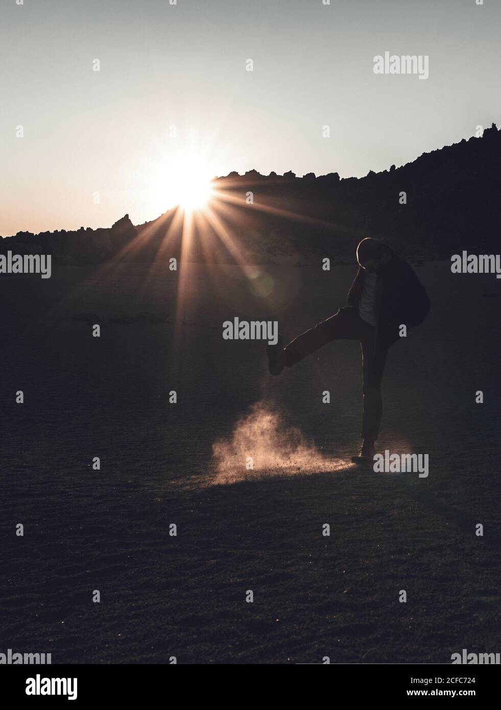 Silhouette of male with lifted leg kicking up sand dust in remote desert standing in sunset back lit on Tenerife Stock Photo