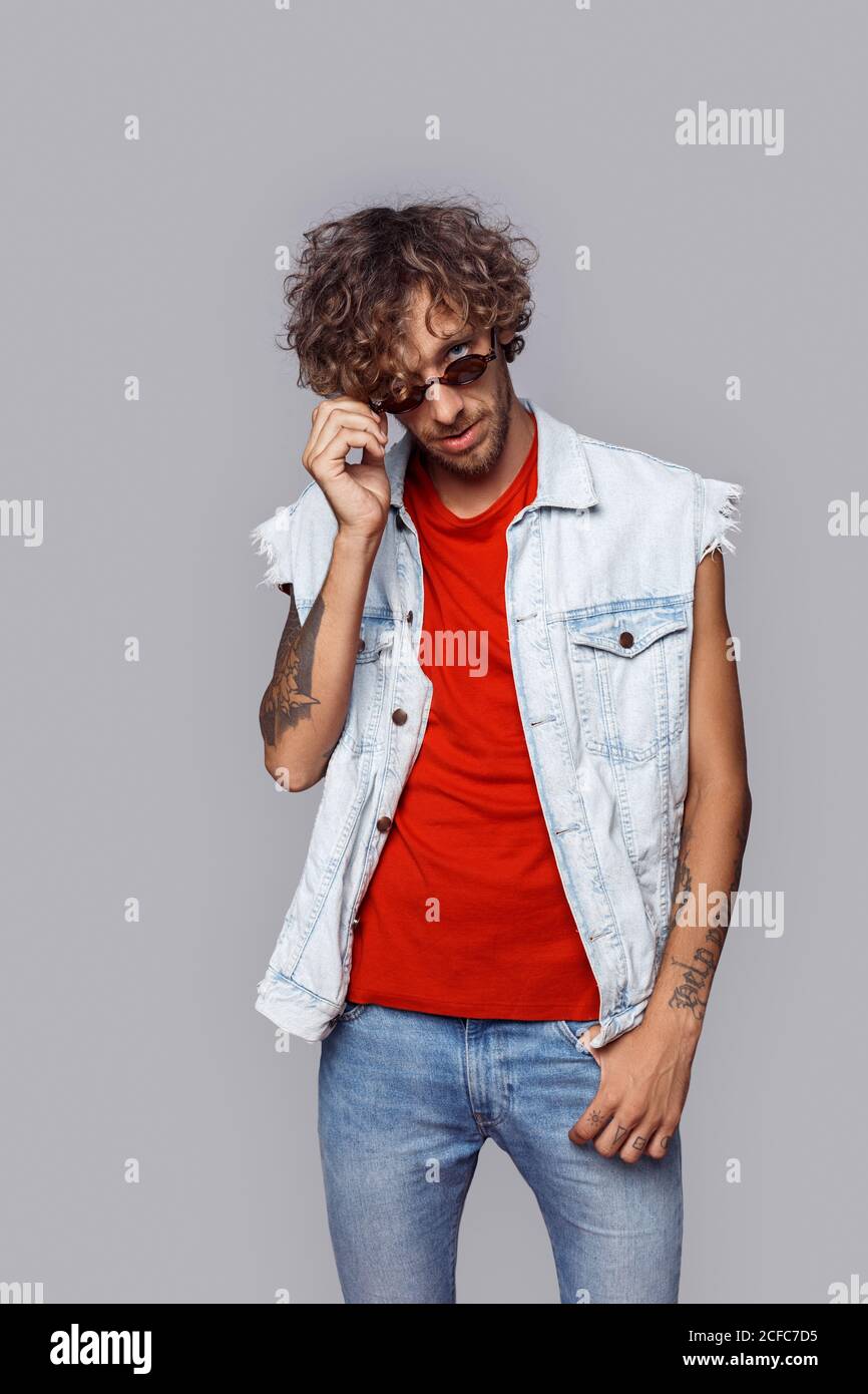 Freestyle. Man with curly hair in jean sleeveless jacket standing isolated on grey taking off sunglasses playful Stock Photo