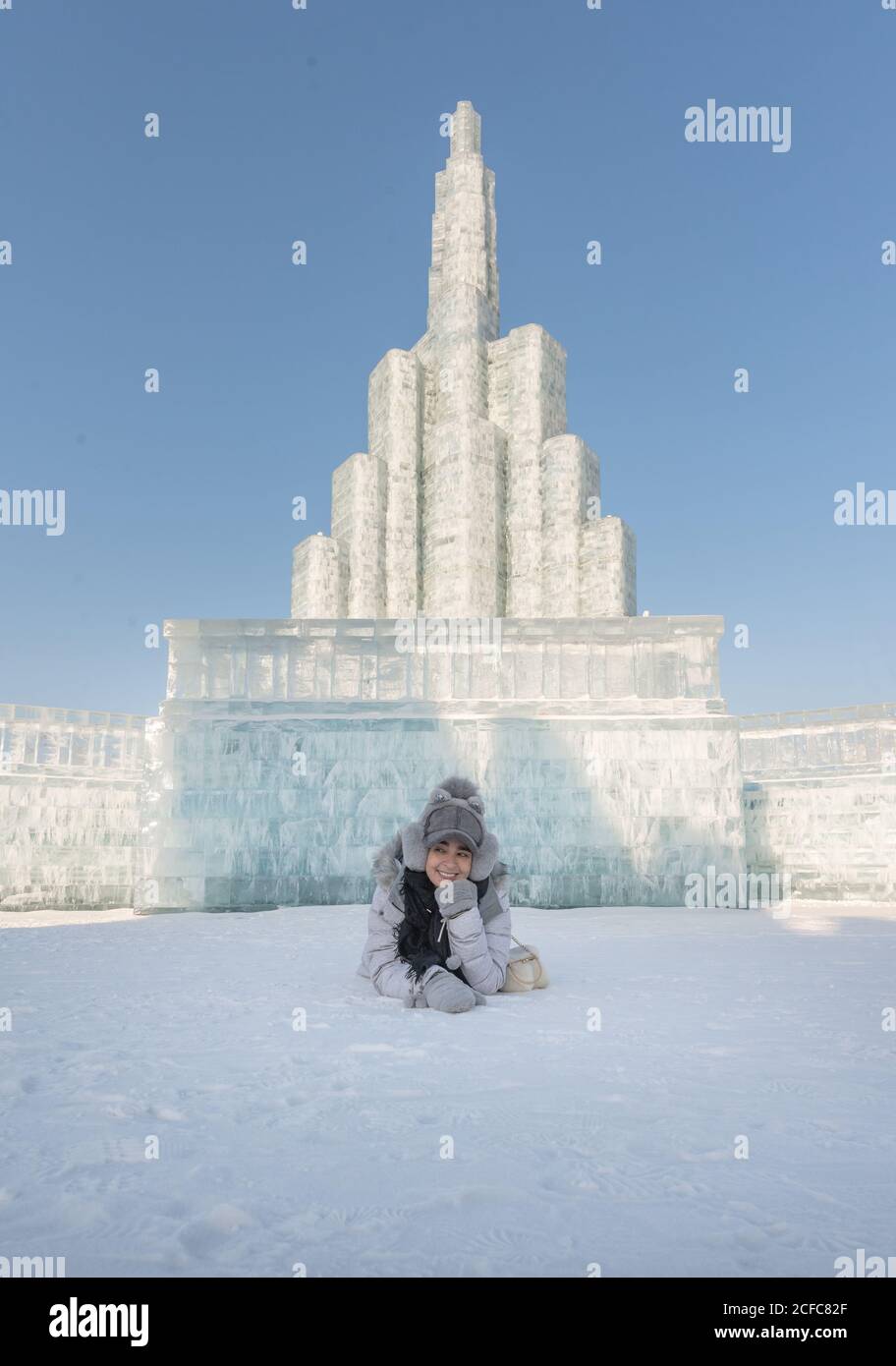 Optimistic Woman in outwear smiling and lying on snow against ice building on cloudless day in winter Stock Photo