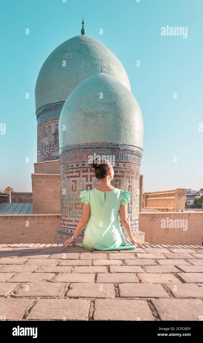 Back view of anonymous Woman sitting on old building against shabby domes of Shah-i-Zinda on sunny day in Samarkand, Uzbekistan Stock Photo