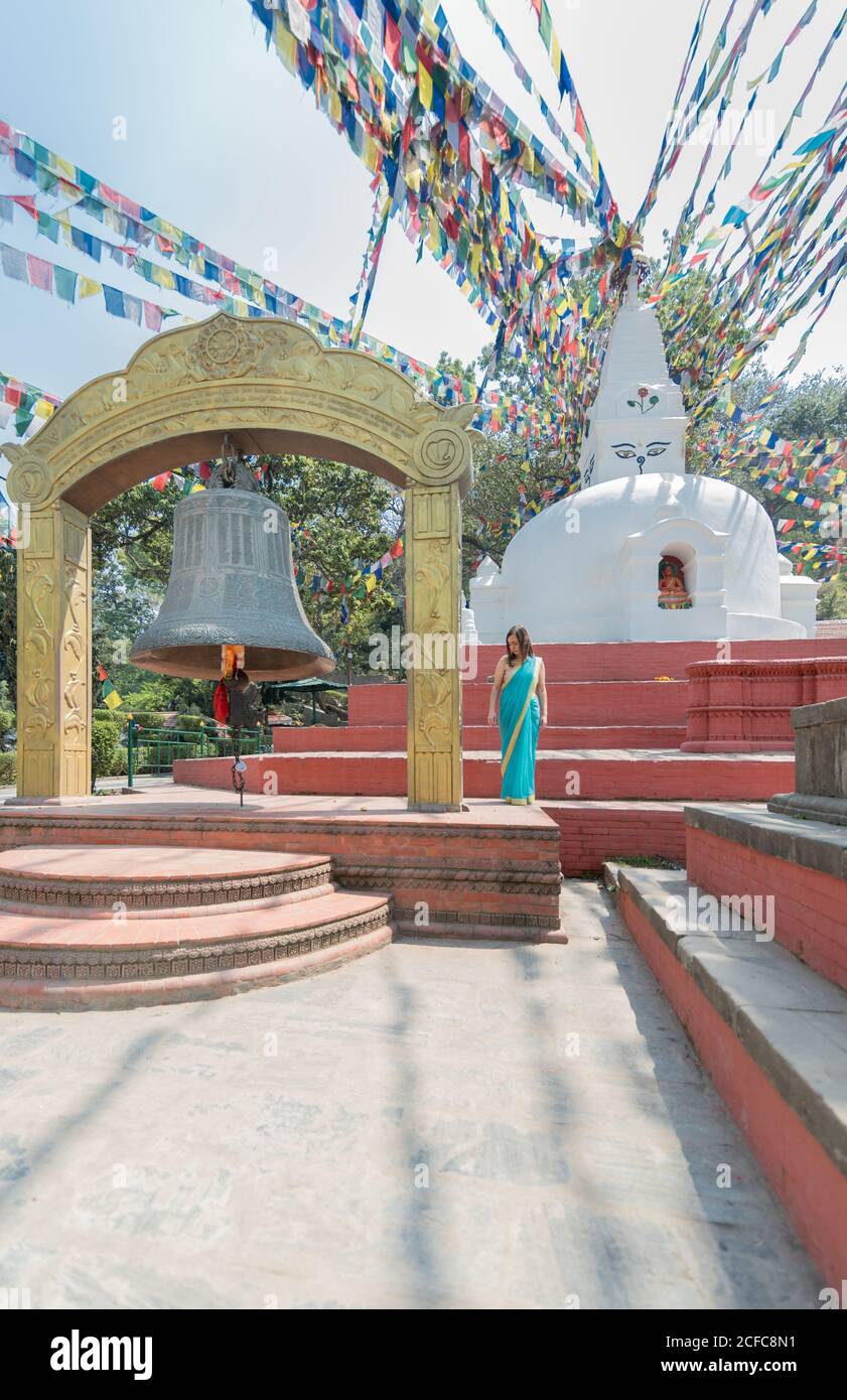 Female tourist in dress looking away while standing on pavement close to old stone hemispherical stupa with cupola on top under garland with flags and big bell in summer Stock Photo