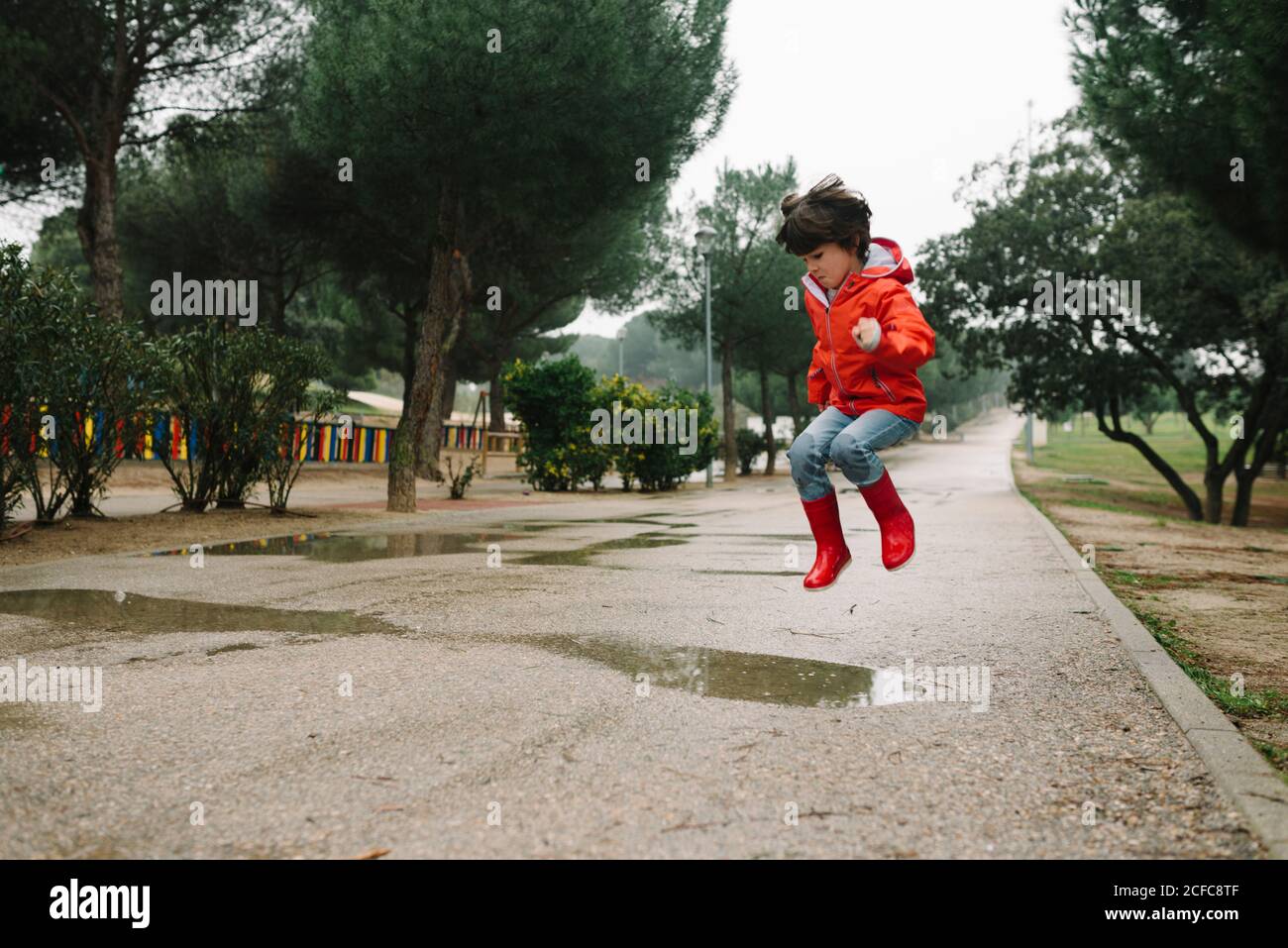 Adorable joyful child in red raincoat and rubber boots having fun jumping  in puddle on street in park in gray day Stock Photo - Alamy