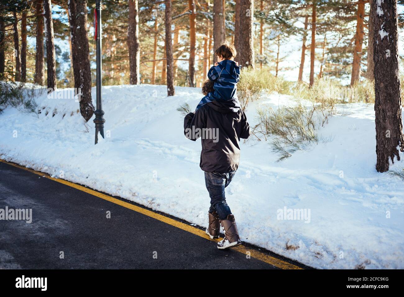 Side view of young man in dark clothes holding child in overall on neck and walking on yellow stripe on road along snowy forest Stock Photo