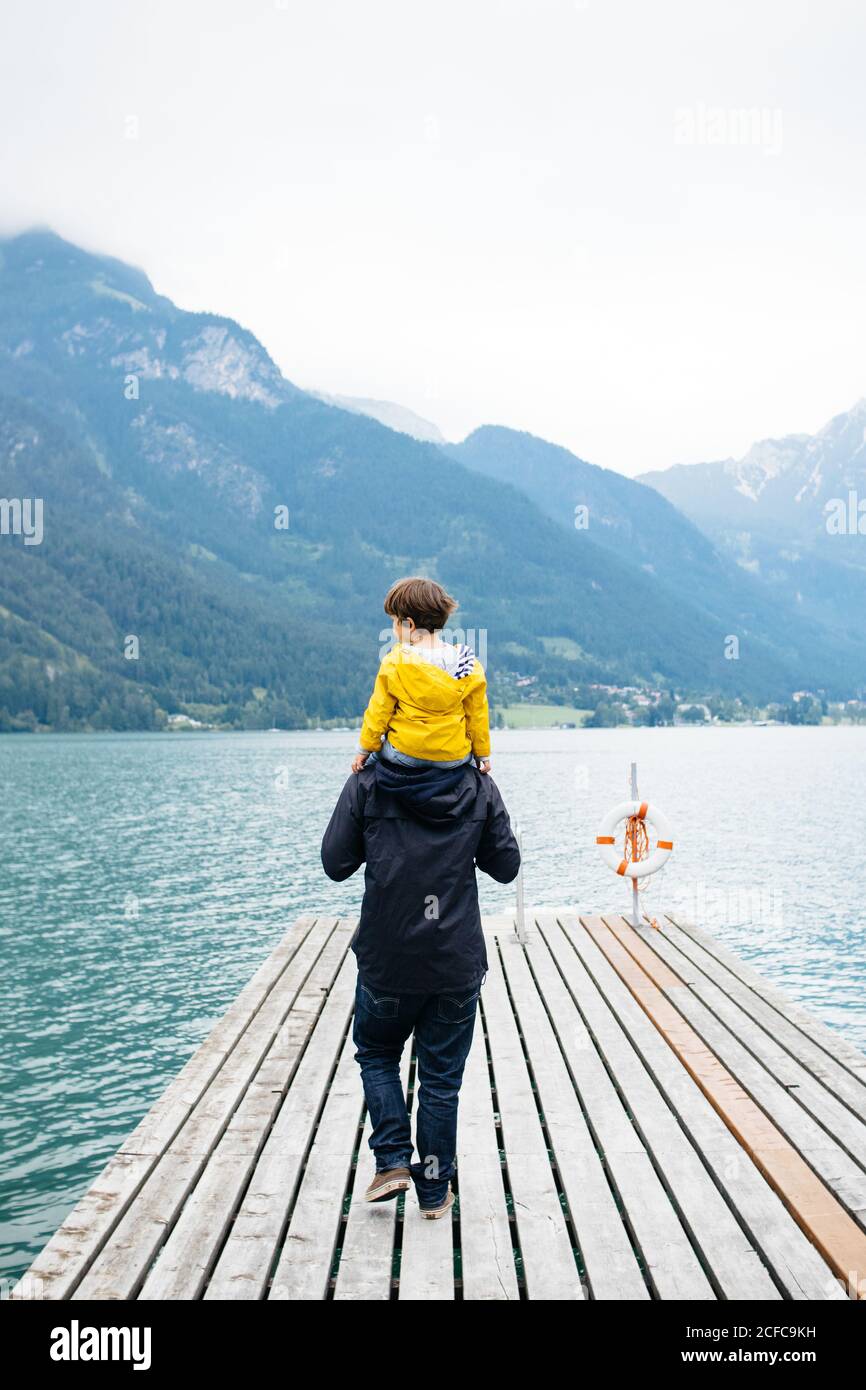 Back view of young man in dark clothes holding child in yellow jacket on neck and walking along wooden pier to calm water and breathtaking landscape in cloudy day Stock Photo