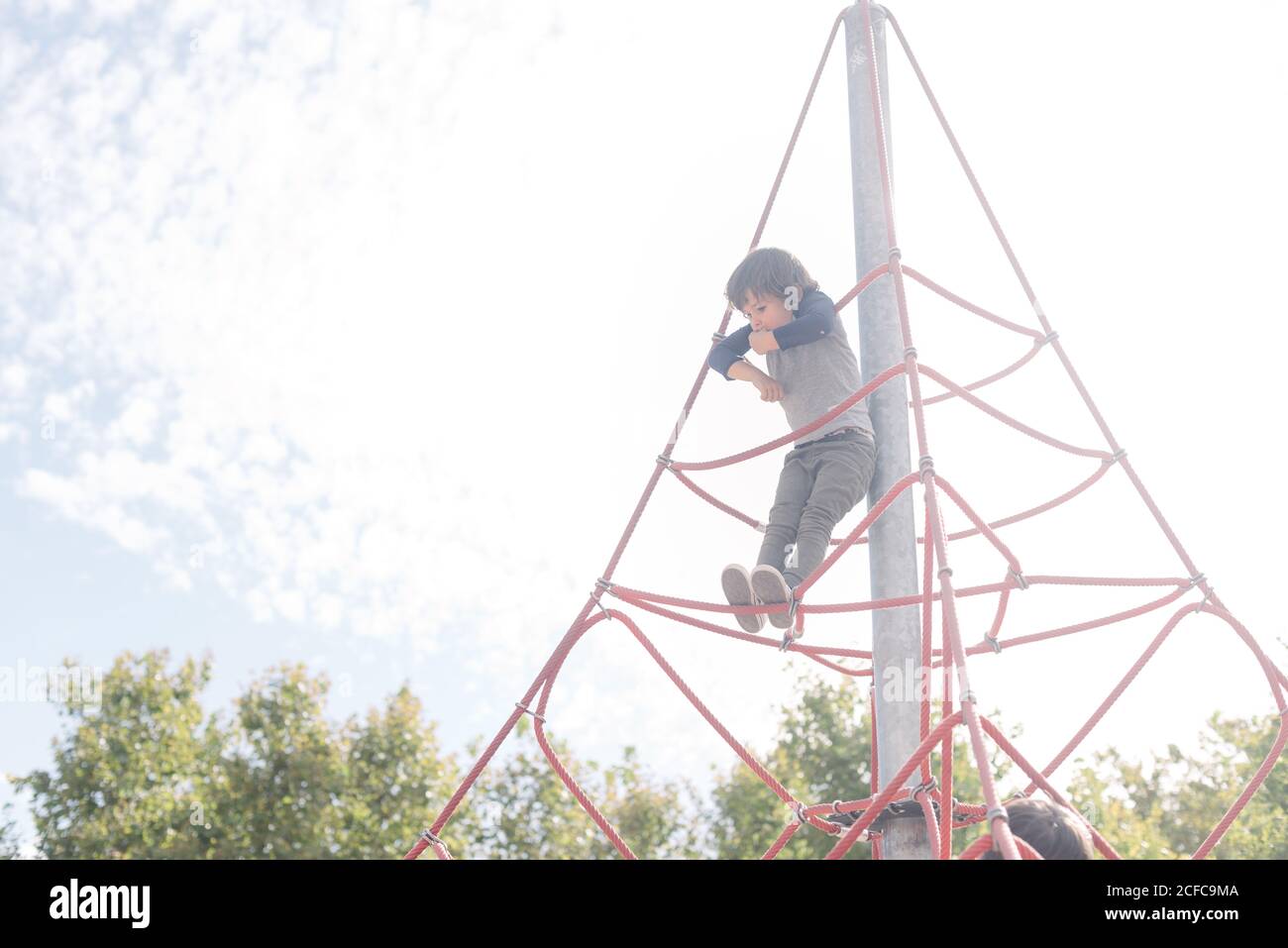 Pensive relaxed kid hanging on rope climbing net on playground in bright light Stock Photo