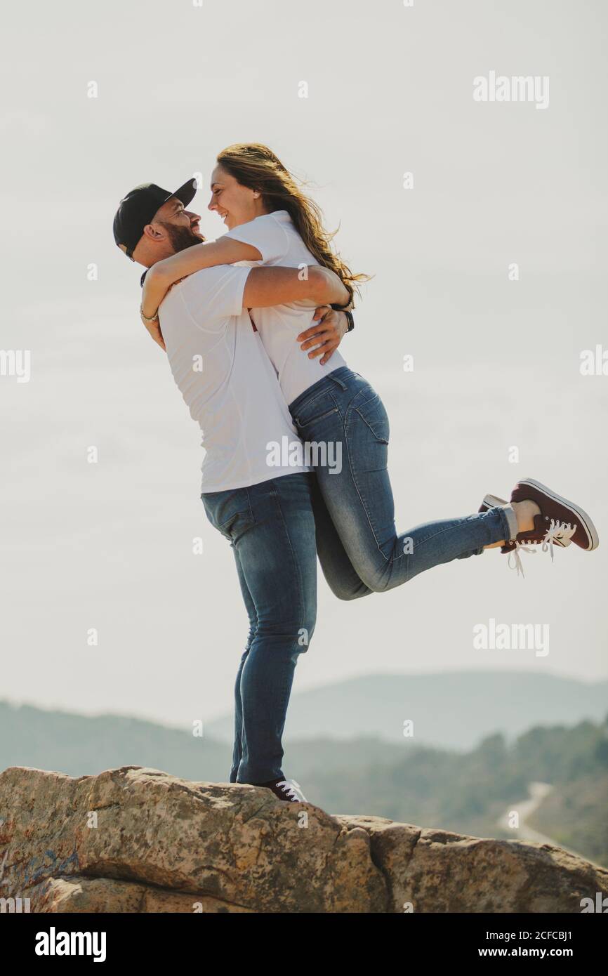 Side view of romantic cheerful couple in matching outfit hugging and looking at each other while standing at hilly slope Stock Photo