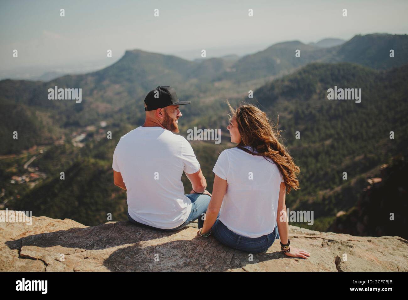 From behind romantic relaxed couple in matching outfit enjoying view while sitting at edge of high cliff in sunlight Stock Photo
