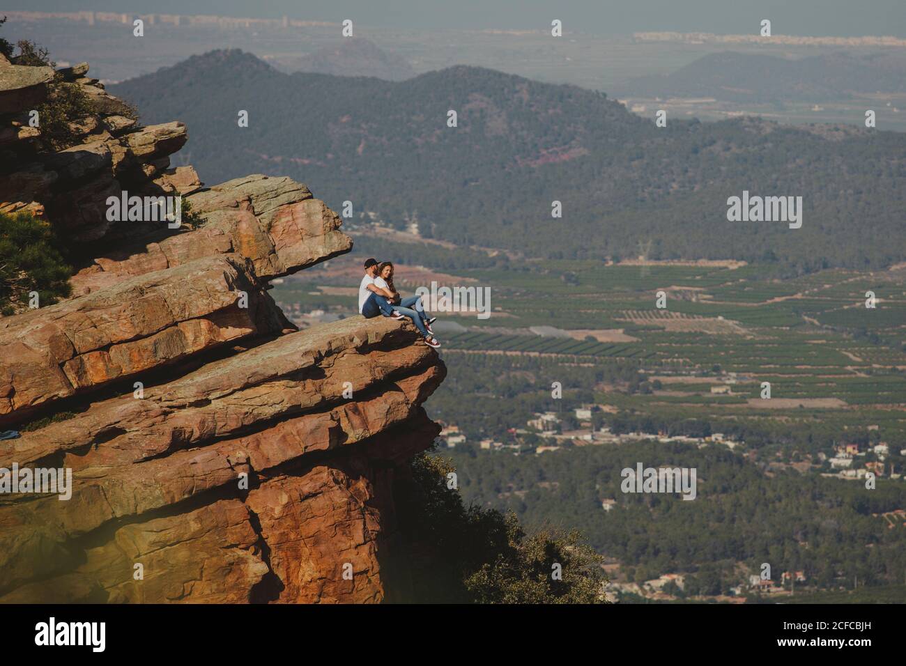 Side view of romantic relaxed couple in matching outfit hugging while sitting on edge of huge rocky mountain Stock Photo