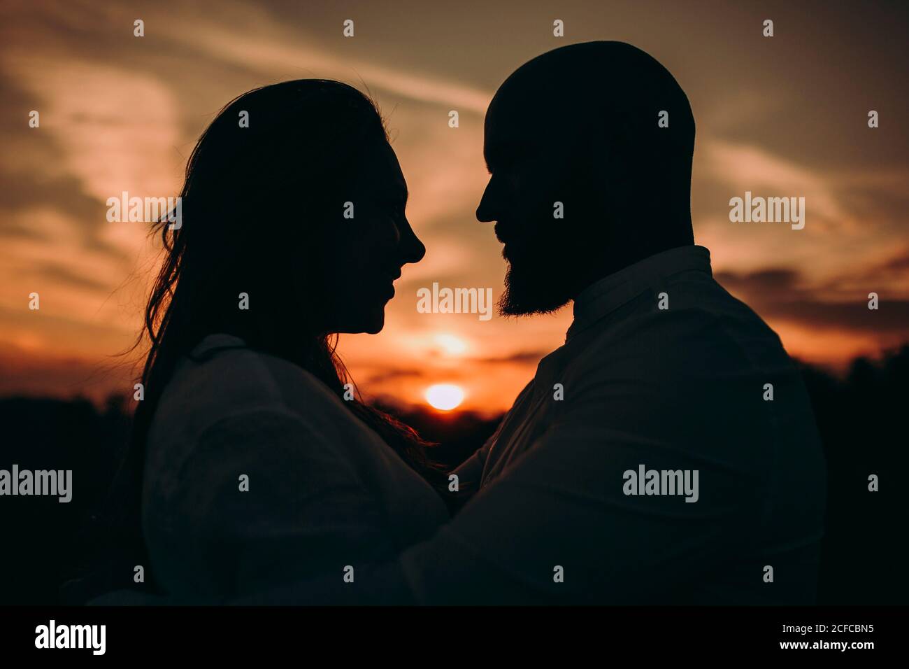 Side view silhouette of caring couple hugging and looking at each other while standing in field in dark red sunset sky Stock Photo