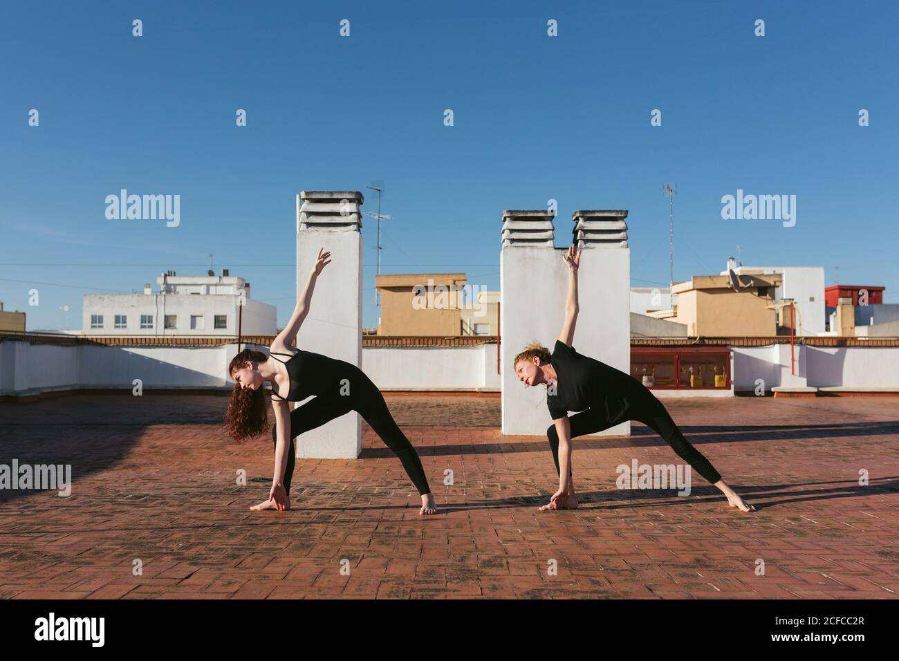 Full body side view of slim women in black activewear performing Revolved Triangle Pose while practicing yoga together on rooftop terrace in city Stock Photo