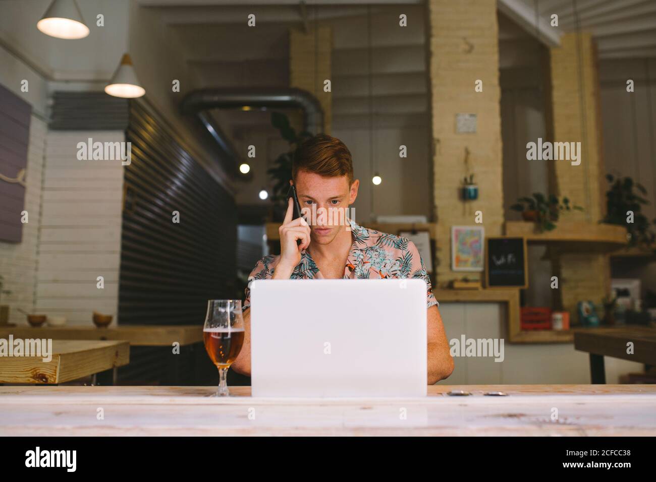Concentrated man talking on smartphone while using laptop at table with drink in kitchen Stock Photo