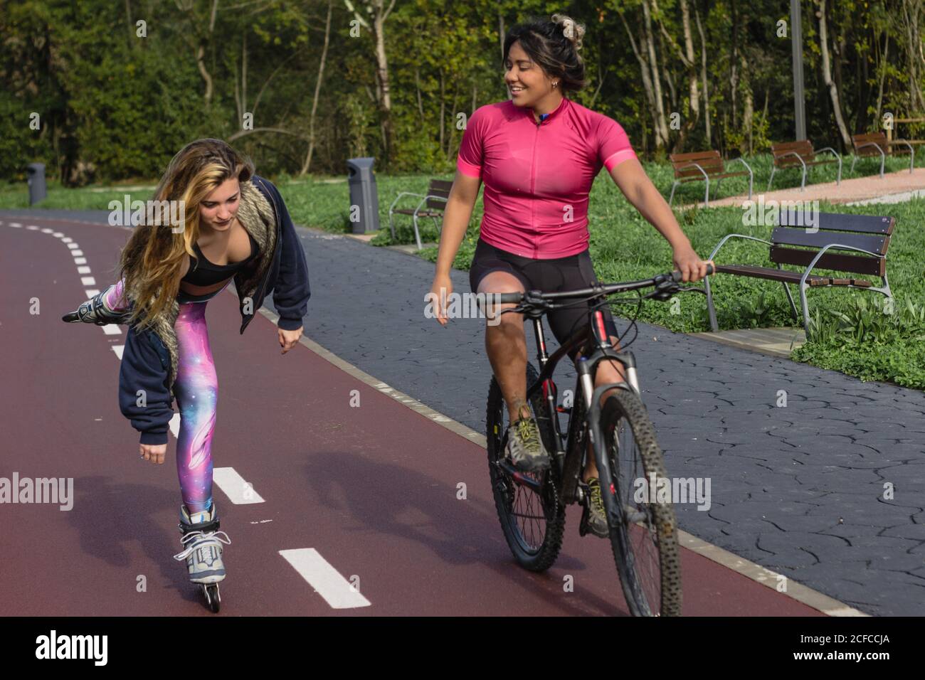 Young multiethnic girlfriends in sportswear actively chatting during weekend while rollerblading and riding bike along bicycle lane in park Stock Photo