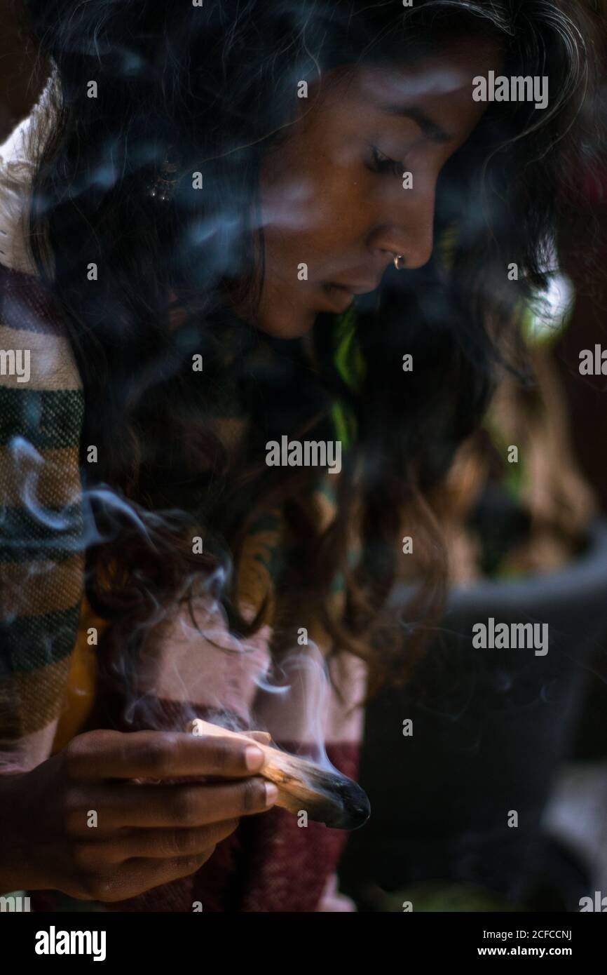 Spiritual young ethnic female with closed eyes in traditional clothes holding burning sage