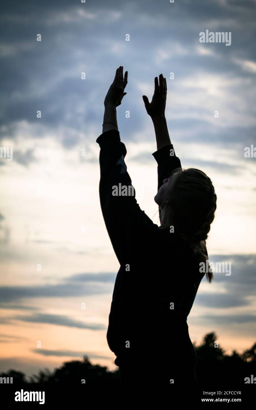 Silhouette of lady raising arms and meditating against sundown sky during workout in countryside Stock Photo