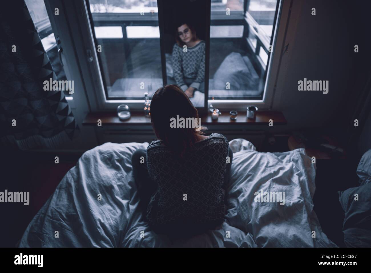 Back view of depressed young female sitting on bed in dark bedroom looking at mirror at home Stock Photo