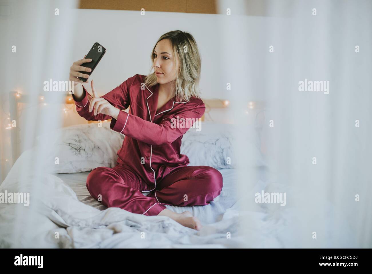 Young pretty Woman in pajamas sitting on bed while doing selfie on ...