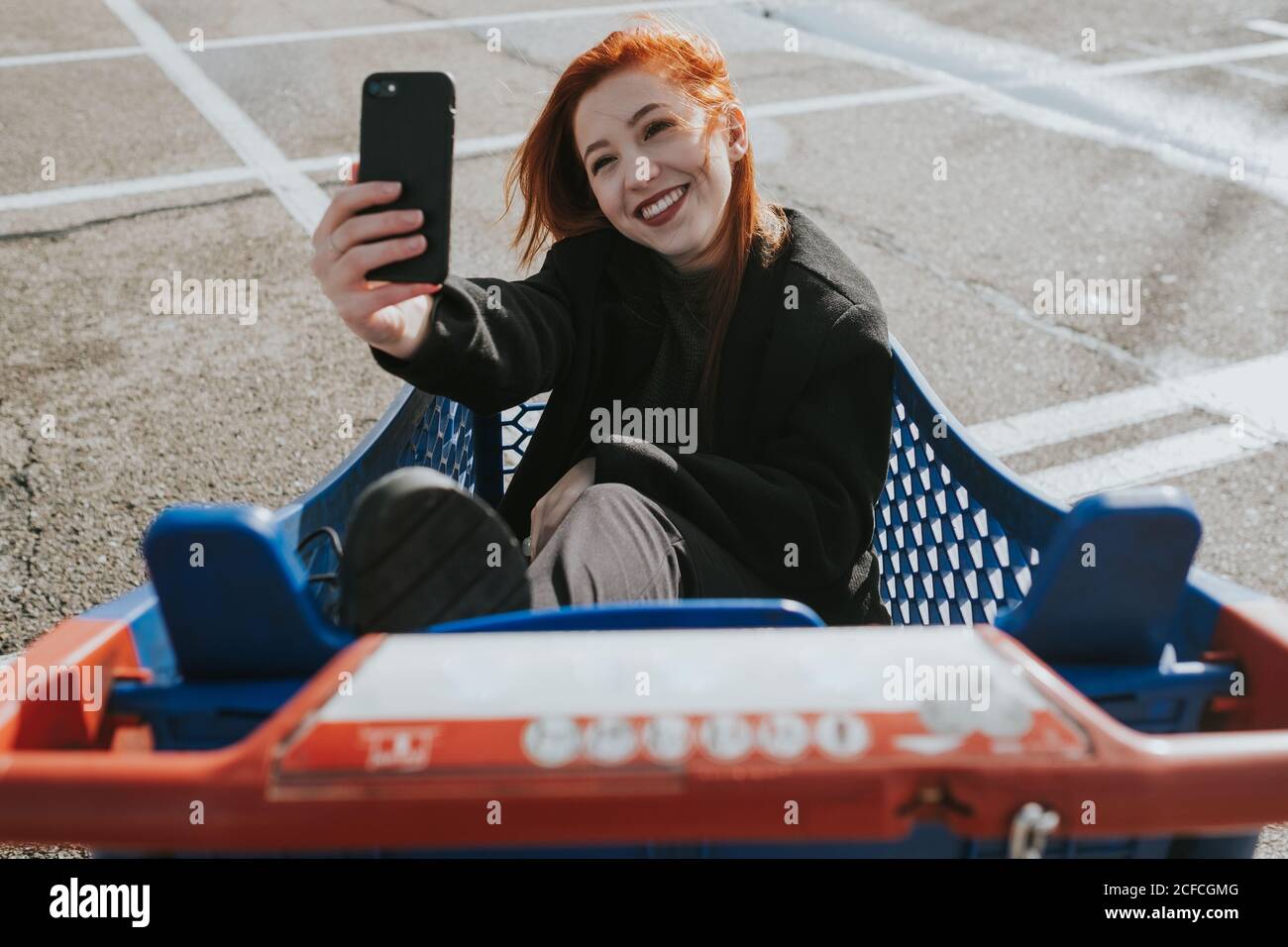 Attractive young Woman with red hair smiling and taking selfie sitting in blue shopping cart with red handle Stock Photo
