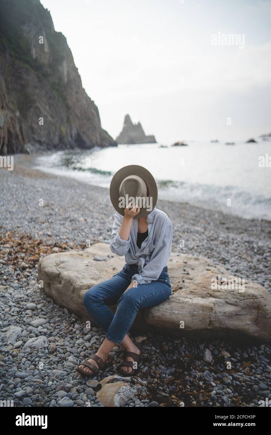 Side view of unrecognizable dreaming stylish female tourist hiding face with hat enjoying seascape while chilling on big stone on rocky shore of Asturias looking away Stock Photo