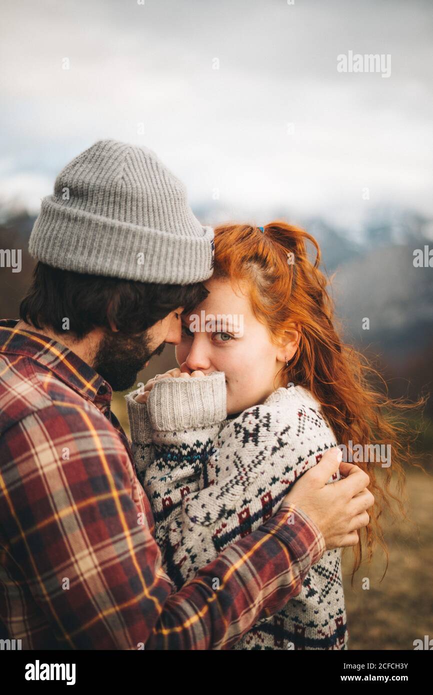 Side view of tender couple hugging and bonding with man closed eyes and Woman looking at camera in cold day in mountains Stock Photo