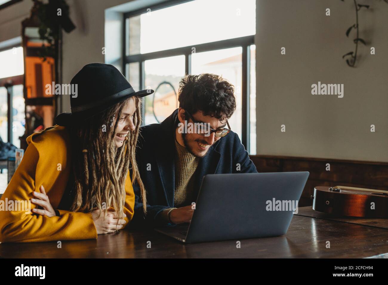 Smart man in glasses and stylish Woman in hat looking at monitor while man typing on laptop in cafe Stock Photo