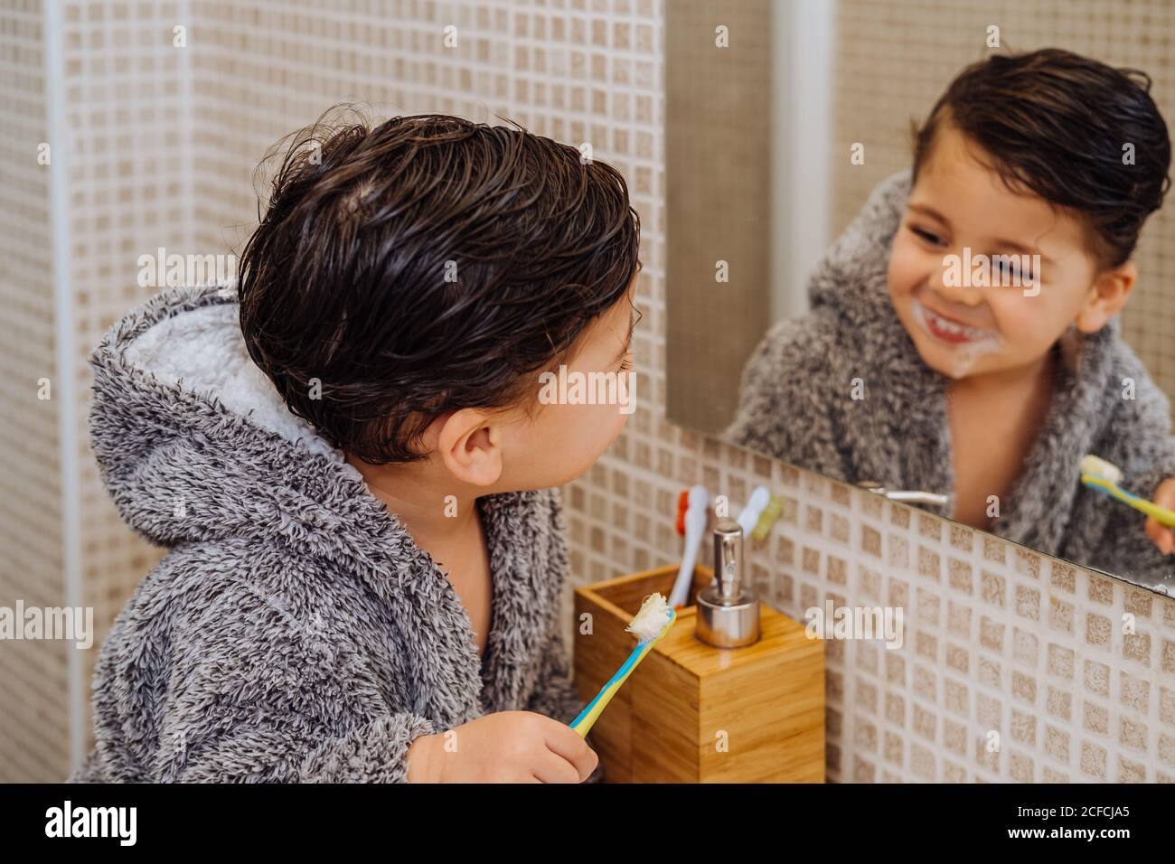 Adorable child wearing cozy bathrobe standing in bathroom with toothbrush and looking in mirror Stock Photo