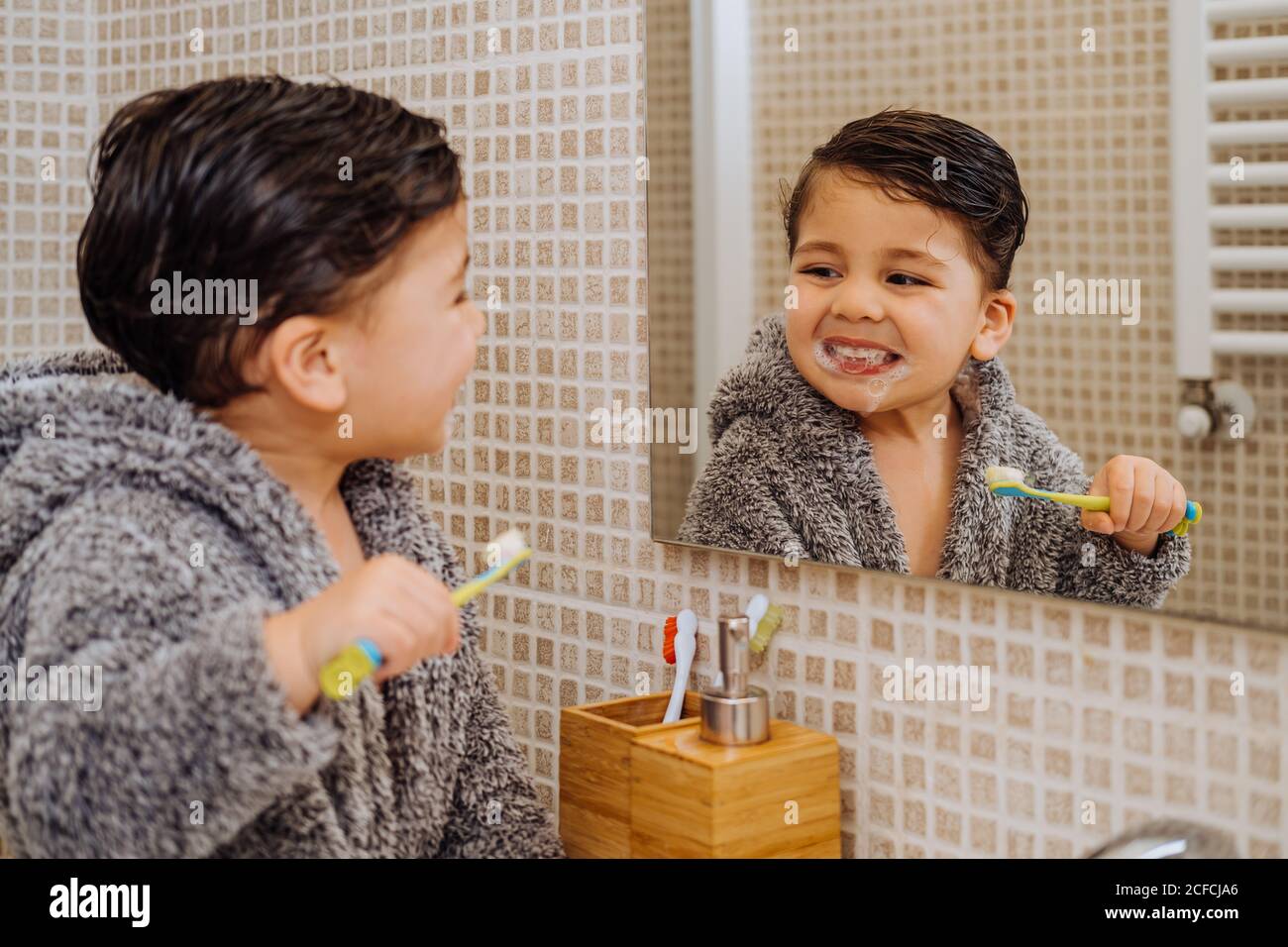 Adorable child wearing cozy bathrobe standing in bathroom with toothbrush and looking in mirror Stock Photo