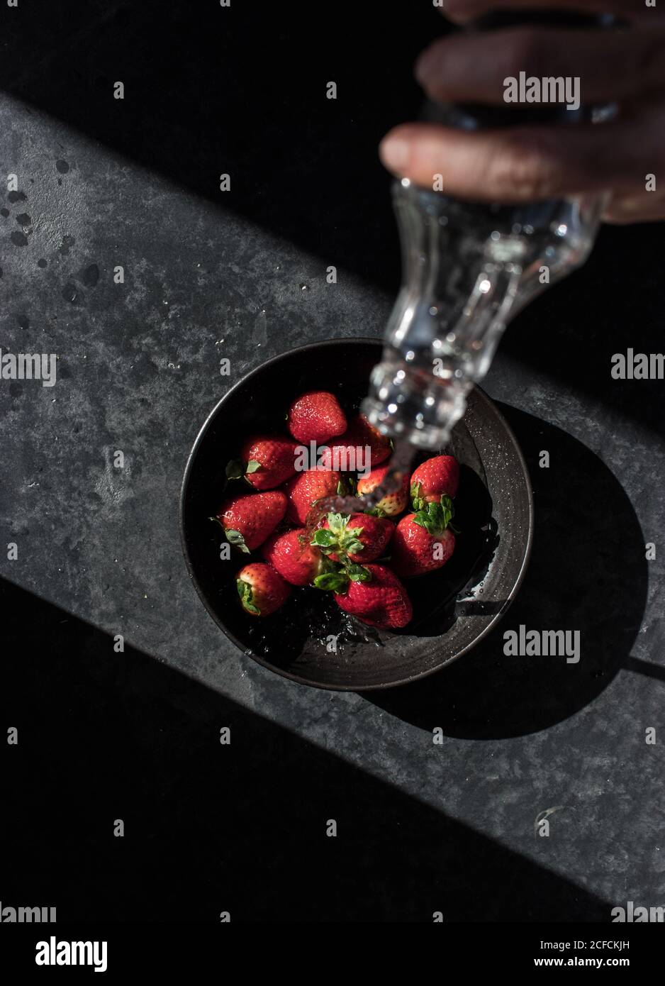 From above crop hand of human holding bottle and pouring liquid of fresh strawberry in bowl illuminated by sunbeams in darkness Stock Photo