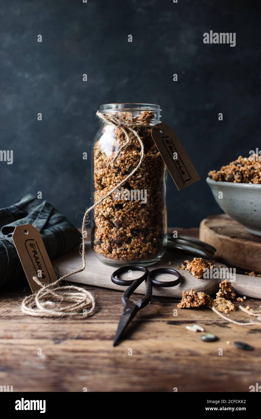Glass jar of fresh millet and quinoa granola with label placed on wooden table near retro scissors Stock Photo
