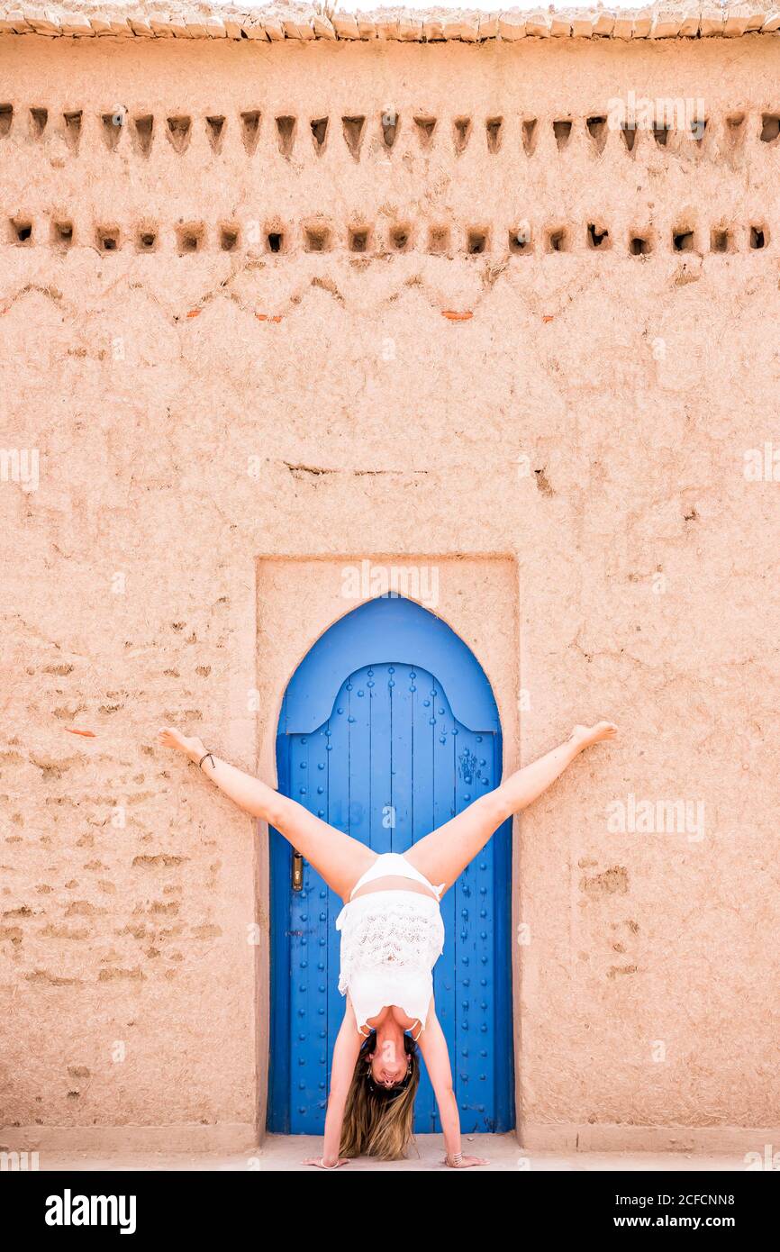 Woman in white beachwear showing handstand against oriental blue door in stone wall, Morocco Stock Photo