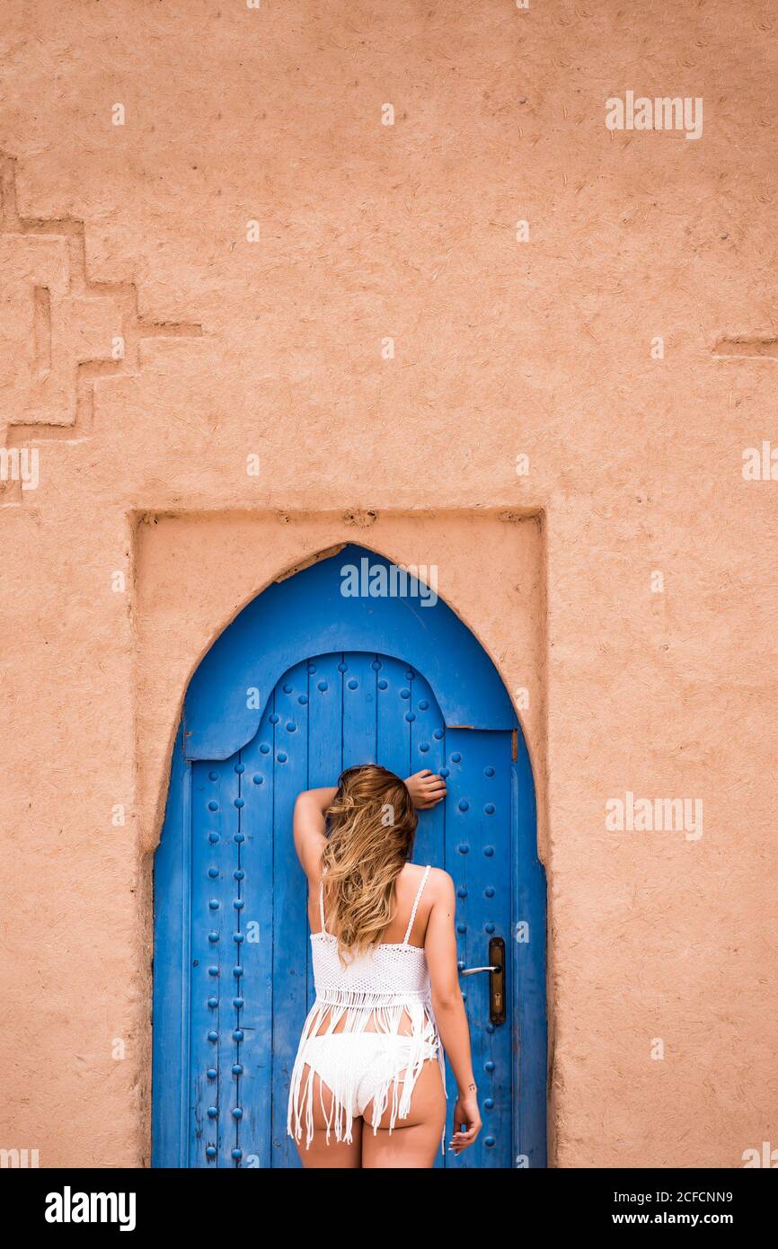 Back view of anonymous Woman wearing white top with bikini leaning against blue oriental door in stone wall, Morocco Stock Photo