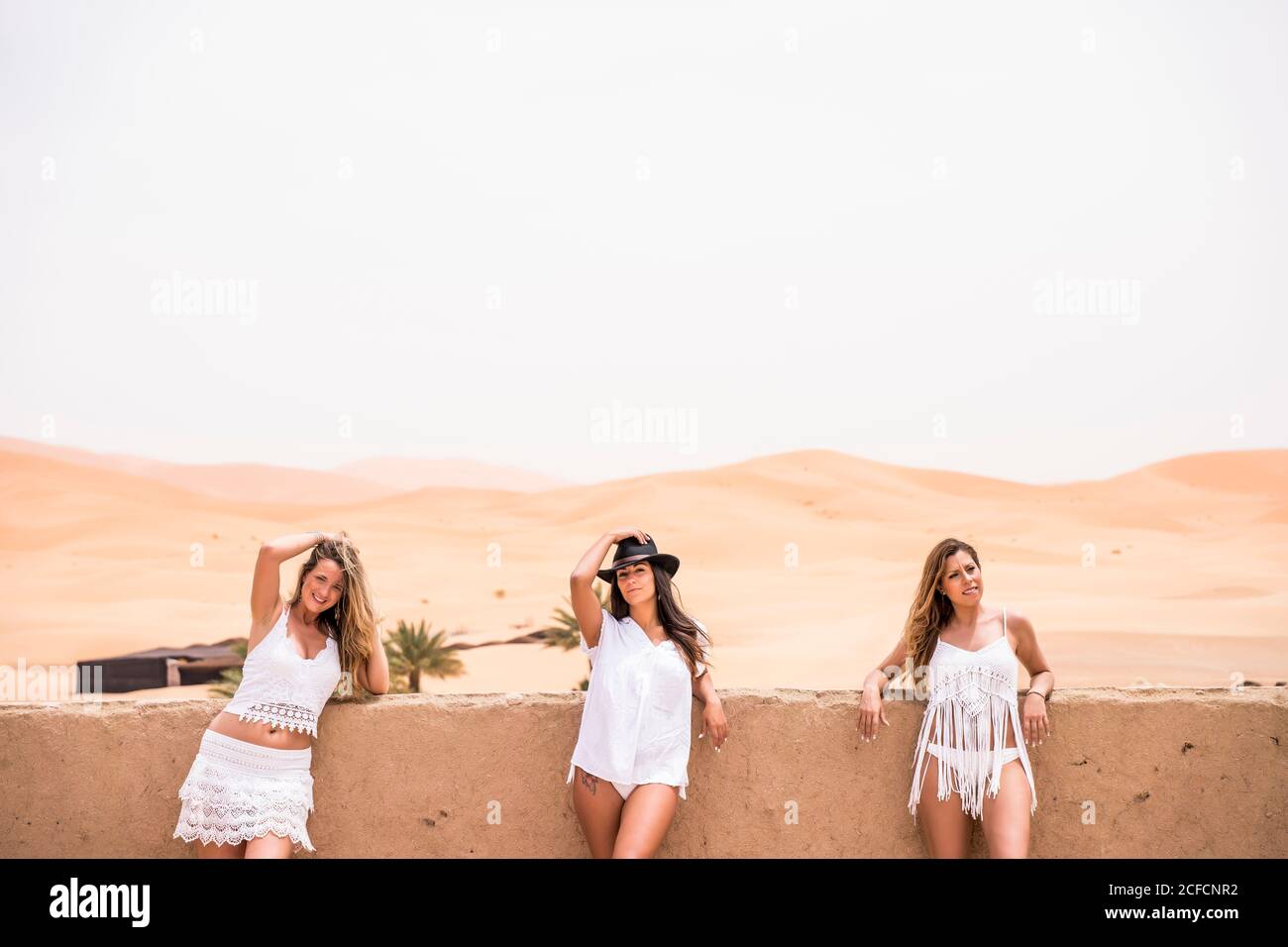Row of women wearing white beachwear posing at stone fence on terrace against endless desert, Morocco Stock Photo