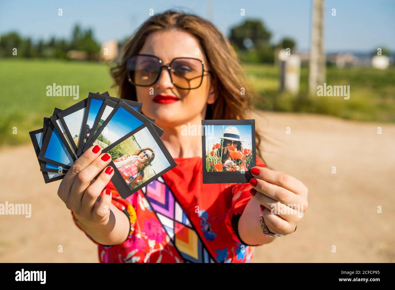 Stylish female demonstrating instant shots with summer memories while standing on blurred background of countryside on sunny day Stock Photo