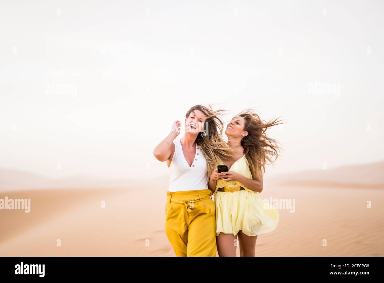 Cheerful stylish two blonde women friends using mobile phone while walking in desert of Morocco Stock Photo