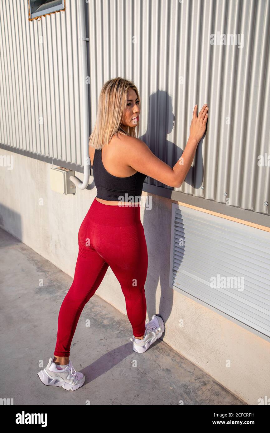 Back view from above of stylish blond haired female in sportive clothes looking away while leaning on wall stretching legs on rooftop Stock Photo