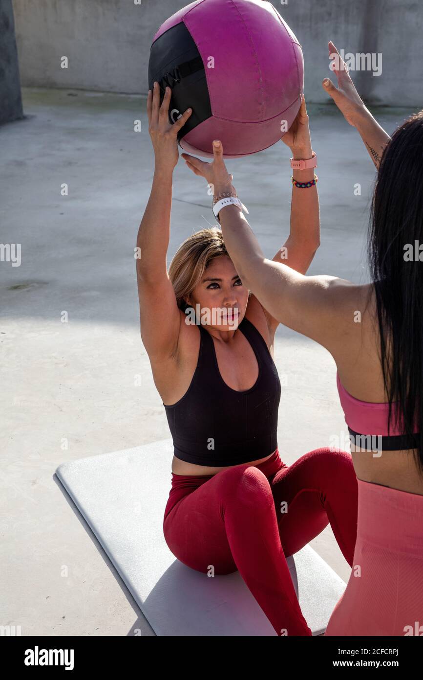 From above of blond haired Woman in stylish active wear sitting on mat with raised hands and passing ball to female sporty partner on rooftop Stock Photo