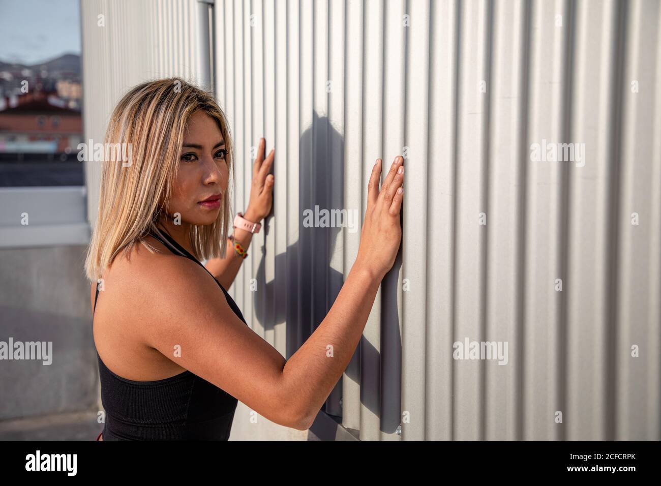 Back view from above of stylish blond haired female in sportive clothes looking away while leaning on wall stretching legs on rooftop Stock Photo