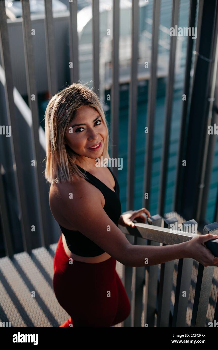 From above of young blond haired lady in black top and red leggings smiling and looking at camera on outdoors stairs Stock Photo