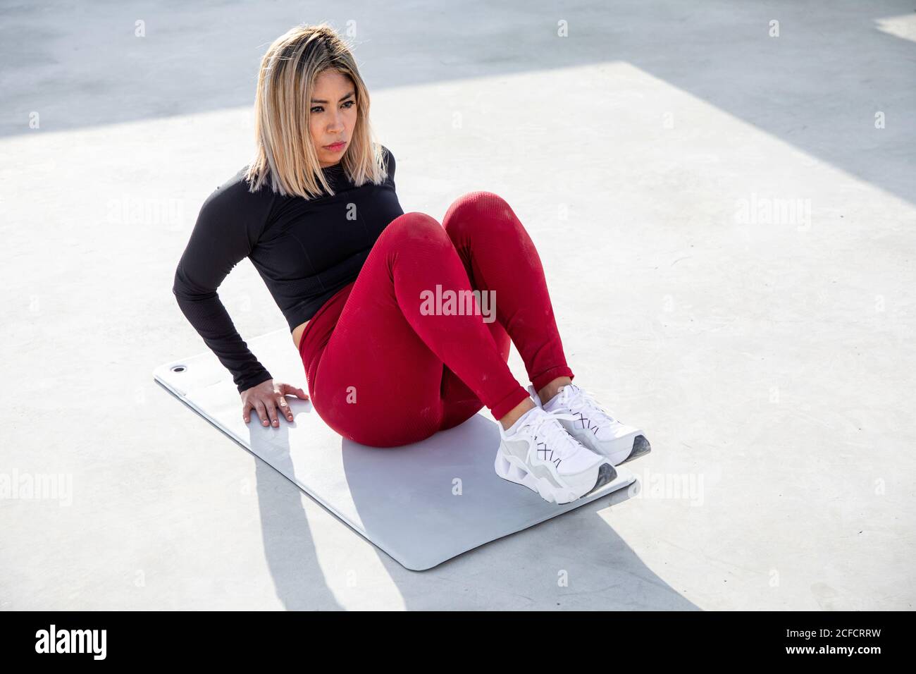 From above of blond haired Woman in sportswear sitting on mat with bent legs training abdomen and looking away on roof of building Stock Photo