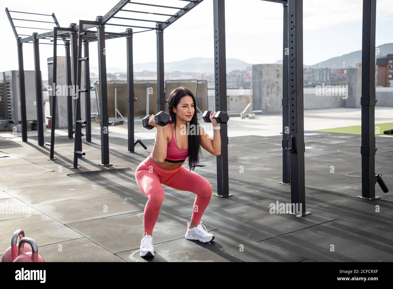 Black haired female athlete in pink active wear in squat position with dumbbells in hands on roof of building looking away Stock Photo