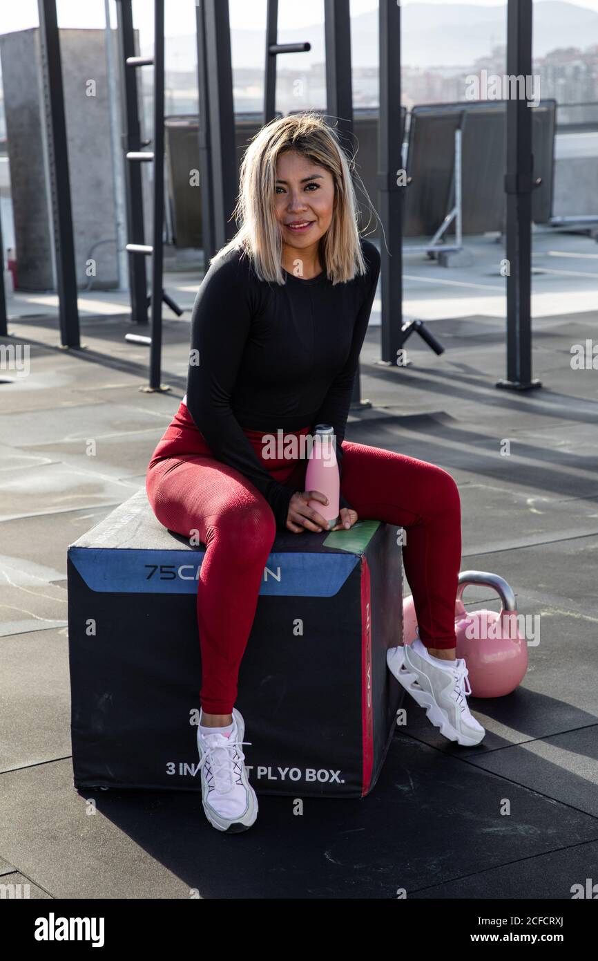 Confident blond haired female athlete in stylish active wear sitting and looking at camera with smile on urban background Stock Photo