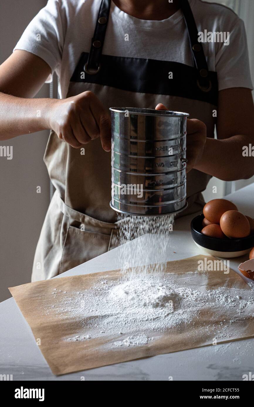 Crop person in apron standing by table sifting flour on baking paper with stainless sifter for