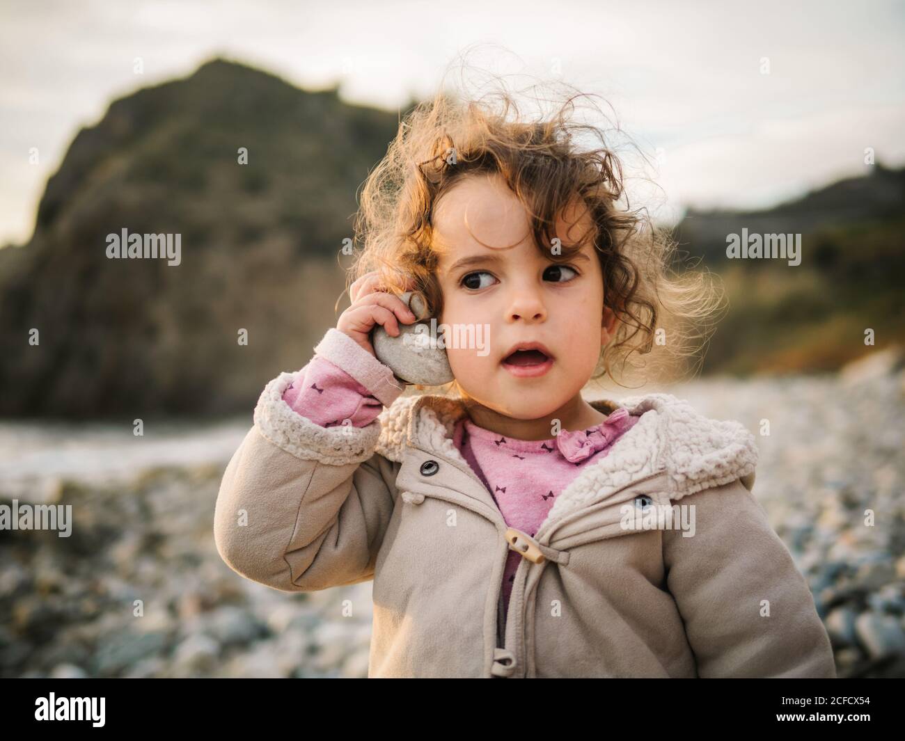 Portrait of cute female kid listening to conch with rapt attention while resting on stony seaside Stock Photo