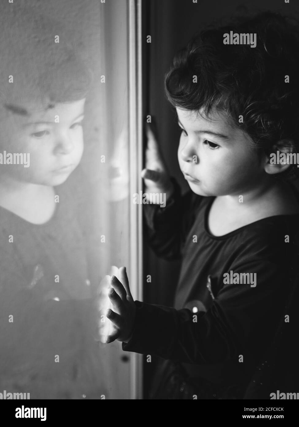black and white portrait of a pensive little boy touching a window with a reflection of himself on it Stock Photo