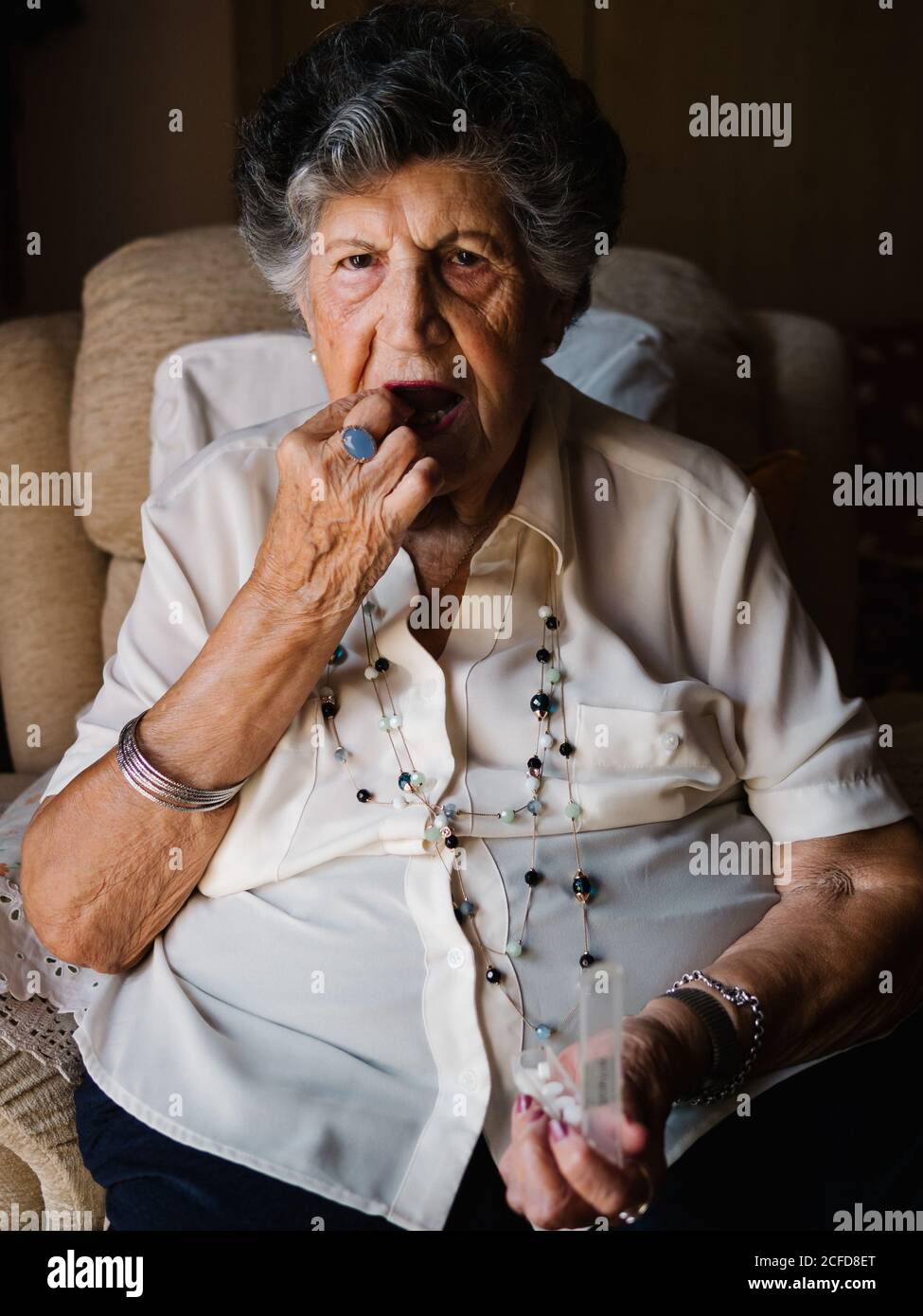 Elegant aged curly gray haired female taking pills sitting on armchair at home and looking at camera Stock Photo