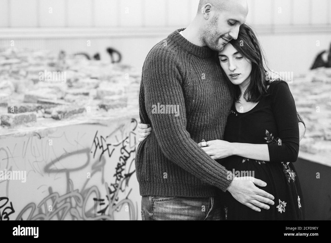 Black and white Side view of bald adult man embracing and kissing pregnant wife while standing on narrow city street together Stock Photo