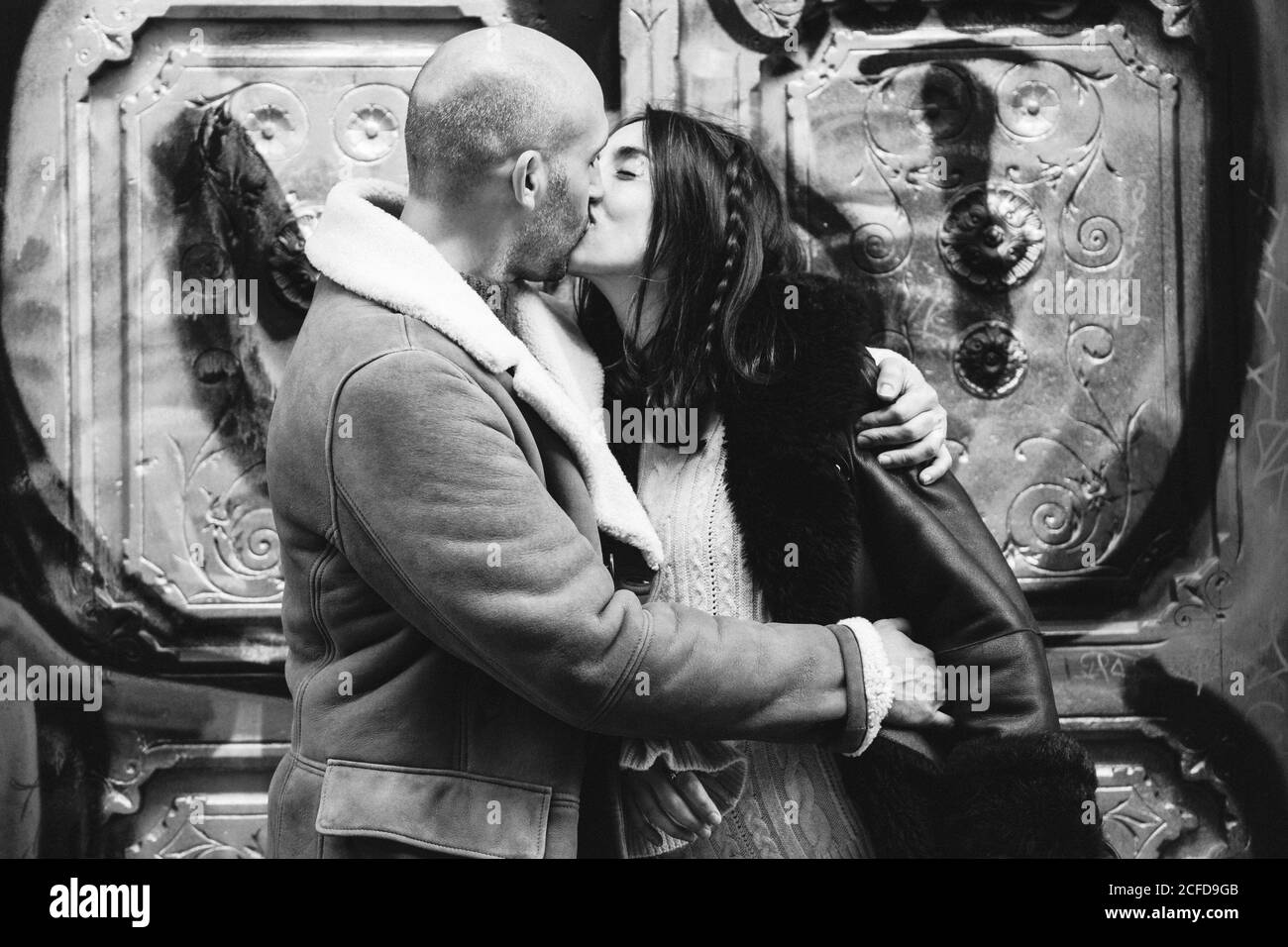 Black and white Adult bald male in coat kissing Woman while standing near graffiti wall on city street Stock Photo