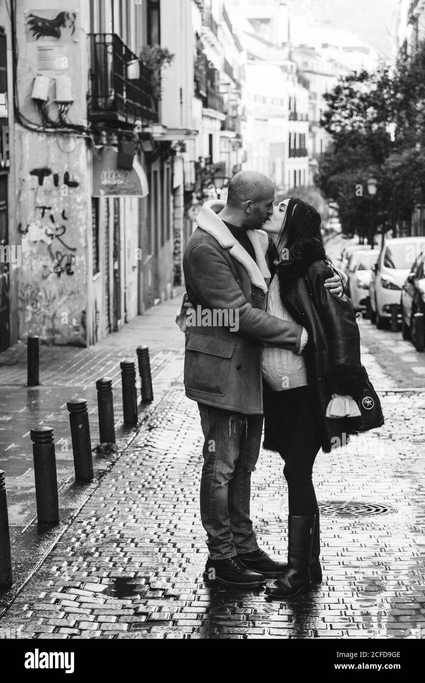 Black and white Side view of bald adult man embracing and kissing pregnant wife while standing on narrow city street together Stock Photo